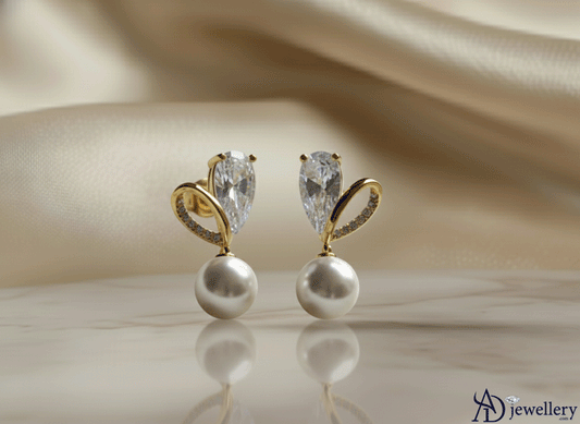 Ethereal pavé leaf and pearl earrings featuring a pear-shaped crystal, gold leaf-inspired design, and dangling white pearl, displayed elegantly on a soft beige fabric background.