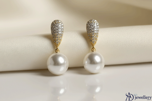A pair of Regal Pearl Glamour Earrings featuring gold teardrop studs encrusted with sparkling pavé diamonds and accented with large, lustrous round pearls, displayed against a soft cream fabric background
